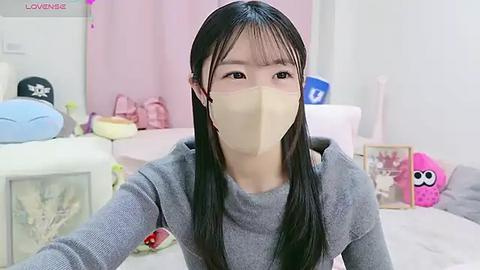 Media: A video of an East Asian woman with long black hair, wearing a beige face mask, grey sweater, and a pink headband, in a soft pink bedroom with stuffed toys and a blue pillow.