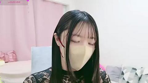 Media: Video of an Asian woman with long black hair, wearing a beige face mask and a black patterned top, indoors with pastel-colored furniture and curtains.