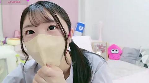 Media: Video of an Asian woman with long black hair, wearing a beige surgical mask, sitting in a pink room with toys and a blue and white \"U\" sign in the background.