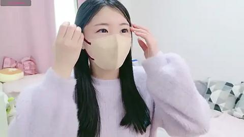 Media: Video of an East Asian woman with long black hair and fair skin, wearing a light purple sweater, beige face mask, and adjusting her glasses. Background features pastel pink curtains, a pink checkered blanket, and a geometric-patterned pillow.