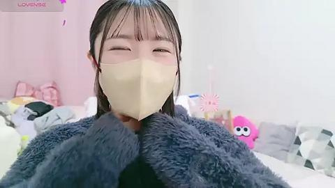 Media: Video of a young Asian woman with straight, black hair, wearing a beige mask and a grey, furry coat, smiling, in a softly lit, pink-themed bedroom with plush toys and a bed.