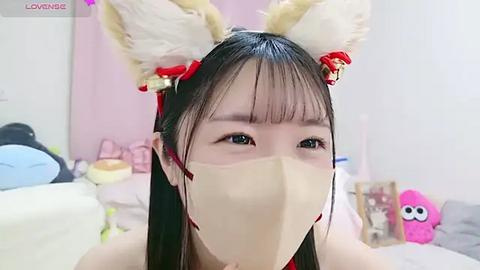 Media: Video of an Asian woman with long black hair and fox ears, wearing a beige face mask, red ribbon, and a pink shirt in a soft, pink-themed room with stuffed toys.