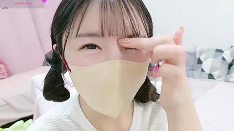 Media: Video of an East Asian woman with dark hair and light skin, wearing a beige mask, white top, and touching her eye. Background includes a bed with white sheets and a checkered pillow.