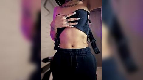 Media: A video of a woman in a dark outfit, lifting her black bra to reveal her bare breasts, with a tattoo on her chest. She wears loose-fitting pants. The background is blurred, suggesting a dimly lit room.