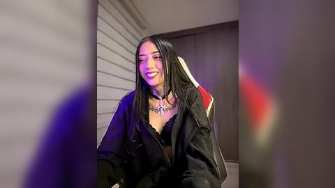 Media: Video of a smiling, medium-toned woman with straight black hair, wearing a black jacket and necklace, seated in a modern, dimly-lit room with white walls and a red and black gaming chair.