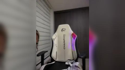 Media: Video of a white, ergonomic gaming chair with a black and red logo on the back, positioned in a modern room with gray walls and a dark wooden door.