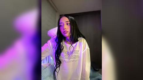 Media: A video of a young woman with long, straight black hair, wearing a white sweater with a bold graphic print, standing indoors, bathed in purple and blue lighting.