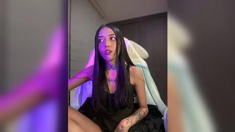 Media: Video of a young woman with long black hair, wearing a black dress, sitting on a bed. She has a tattoo on her right arm and a necklace. The background shows a blurred bedroom with purple lighting.