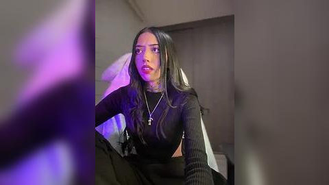 Media: Video of a young woman with long dark hair, wearing a black sweater and black pants, sitting on a bed in a dimly lit room.