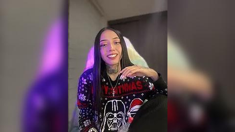 Media: Video of a young woman with light brown skin and long, straight hair, wearing a black \"Star Wars\" Christmas sweater, smiling, in a dimly lit room with purple and green light effects.