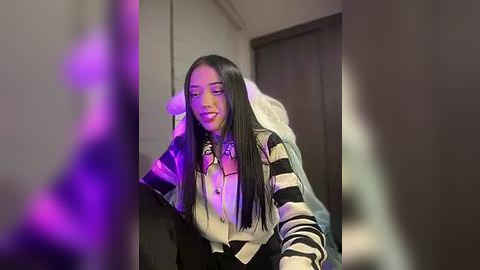 Media: A video of a young woman with long, straight black hair, wearing a striped sweater and a black skirt, smiling. The background shows a dimly lit room with purple lighting.