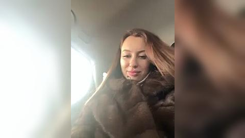 Media: A video of a young Caucasian woman with long, light brown hair and fair skin, wearing a dark fur coat, seated in a plane with a window beside her. The image is slightly blurry.