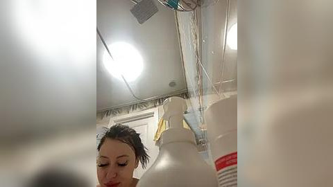 Media: A video of a woman with dark hair in a ponytail, wearing a white shirt, holding a white jug, in a bright, sterile hospital room with medical equipment and a white ceiling light.