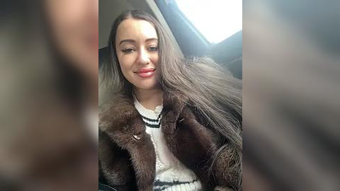 Media: Video of a young woman with long brown hair, fair skin, and red lipstick, wearing a white sweater and brown fur coat, seated in a car with daylight filtering through the window.