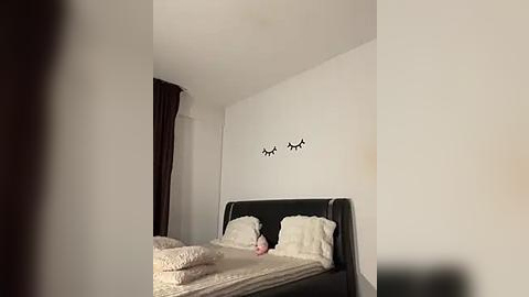 Media: A video of a minimalist, modern bedroom featuring a black bed with white bedding, a plush pillow, and a pink toy. White walls with two black, eyelash-shaped wall decals. Dark curtains and a clean, uncluttered space.