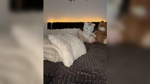 Media: Video of a cozy bedroom with a white, fluffy throw blanket on a dark, textured bedspread. Two stuffed animals, a white bunny and a brown bear, sit against a wall lit by warm, string lights.