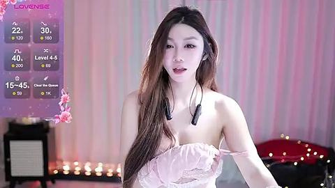 Media: Video of an East Asian woman with long brown hair, wearing a strapless pink dress, standing in a dimly lit room with pastel-striped wallpaper.