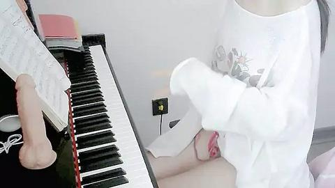 Media: Video of a person in a white long-sleeve shirt playing a piano, with a partially visible, fair-skinned person lying on their back, exposing their genitals. The setting appears to be a bright, possibly clinical environment.