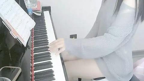 Media: A video of a person with long black hair, wearing a light gray sweater, playing a black piano. The background features a white wall and scattered papers, creating a casual, homey atmosphere.