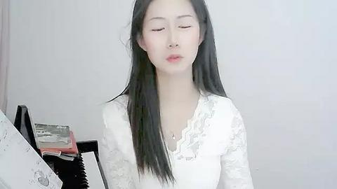 Media: Video of a serene Asian woman with long black hair, wearing a white lace dress, sitting at a piano, playing softly, in a minimalist room.