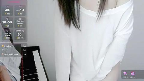 Media: Video of a woman with long black hair, wearing a white off-shoulder top, standing in a simple room with a digital piano in the background.