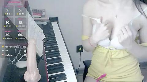 Media: Video of a young woman with fair skin and small breasts, wearing a yellow skirt, sitting at a piano. A realistic, white dildo is on the piano.