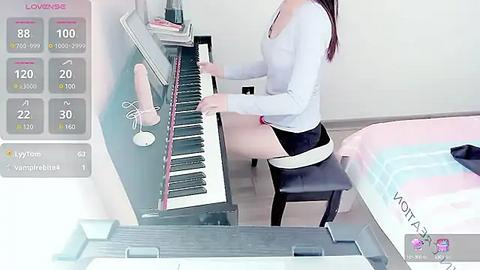 Media: Video of an Asian woman with long brown hair, playing a digital piano in a modern bedroom, wearing a light grey top and black skirt.