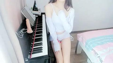 Media: Video of an East Asian woman in a white crop top and pink panties, seated at a black piano in a brightly lit bedroom.