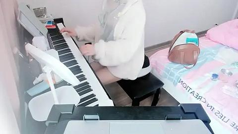 Media: A video captures a person with a white fur hood playing a digital keyboard in a minimalist room with a pink bedspread, white walls, and a brown handbag.