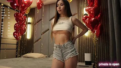 Media: Video of a slender, light-skinned Asian woman with long brown hair in a white crop top and denim shorts, surrounded by red balloons in a modern bedroom.