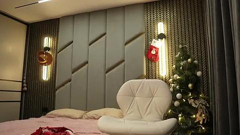 Media: Video of a modern, cozy bedroom with a gray, geometric-patterned headboard, white leather chair, Christmas tree adorned with ornaments, and warm ambient lighting.