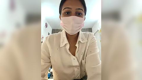 Media: Video of a South Asian woman with medium brown skin, wearing a white button-up shirt, pink surgical mask, and dark hair tied back. Background shows a medical setting with blurry white walls and medical equipment.