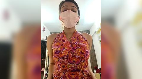 Media: Video of a young woman with light brown skin, wearing a pink surgical mask, a colorful, halter-neck dress, and a headscarf, indoors in a blurred, well-lit room.