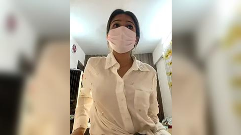 Media: Video of a woman with medium brown skin and dark hair, wearing a white button-down shirt, pink face mask, and a white surgical gown, standing indoors, blurred background.