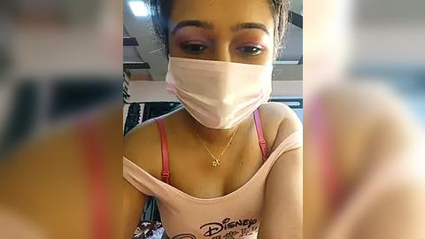 Media: Video of a young woman with light brown skin, wearing a pink Disney-themed tank top, a face mask, and a gold necklace, sitting in a car with blurred background.