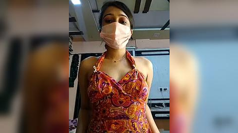 Media: Video of a young woman with medium brown skin, dark hair tied back, wearing a pink surgical mask, a colorful paisley-patterned apron, and a sleeveless top. She stands indoors with a blurred background, possibly a kitchen.