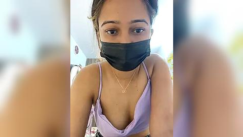 Media: Video of a young woman with light brown skin, wearing a black face mask, purple bralette, and gold necklace, sitting indoors with blurred background.