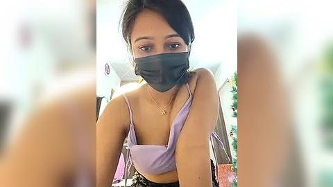 Media: Video of an Asian woman with medium skin tone, wearing a black face mask, a light lavender crop top, and a black skirt. Background is blurred, showing indoor setting with shelves and greenery.