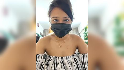 Media: Video of a young Asian woman with medium brown skin, wearing a black face mask, black off-shoulder top, and gold necklace, in a blurred indoor setting.