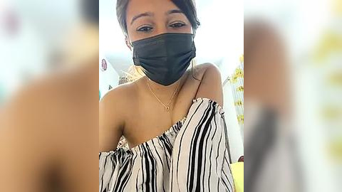 Media: Video of a young woman with medium brown skin, wearing a black face mask, off-shoulder black and white striped top, and a delicate necklace. Background is blurred, featuring a yellow wall and a person.