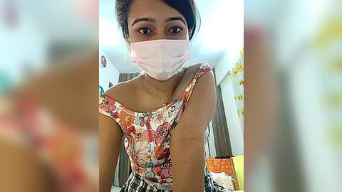 Media: Video of a young South Asian woman with a medium brown complexion, wearing a floral-patterned sleeveless top and a pink face mask, seated indoors with a blurred background.