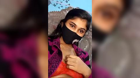 Media: Video of a young woman with medium-brown skin, wearing a black mask and a pink and black striped jacket, sitting on a grey couch, with blurred background.