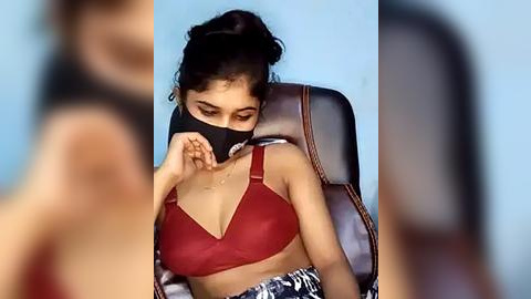 Media: Video of a young woman in a red bra, wearing a black mask and a patterned skirt, seated in a leather chair against a light blue wall.