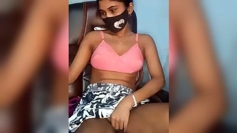 Media: Video of a young woman with dark skin, wearing a pink bra, patterned skirt, and face mask, seated in a chair against a blue wall.