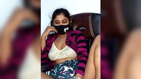 Media: A video of a woman wearing a black face mask, striped cardigan, and floral skirt, seated in a dark leather chair.