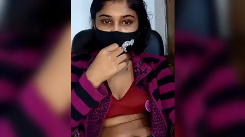 Media: Video of a woman with medium brown skin, wearing a black face mask, red bra, and purple-striped cardigan, sitting indoors with a blurred background.