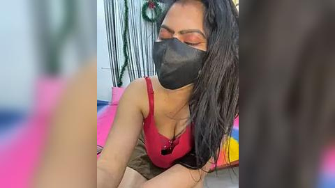 Media: Video of a woman with long black hair, wearing a red tank top, black face mask, and brown pants, lying on a colorful blanket in a room with festive decorations.