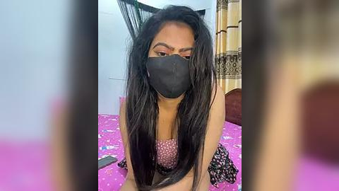 Media: Video of a South Asian woman with long black hair, wearing a black face mask, pink top, and polka-dot skirt, sitting on a pink bed with a patterned sheet, in a room with a beige curtain and a wooden headboard.