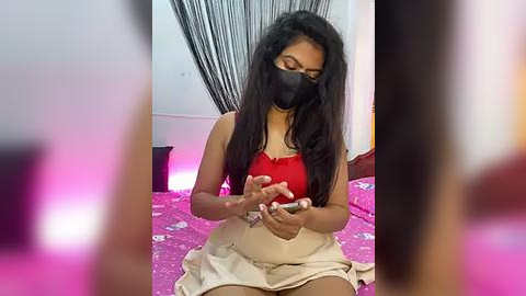 Media: A video of a South Asian woman with long black hair, wearing a red crop top and beige skirt, playing with a smartphone on a pink bedspread.