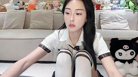 Media: Video of an East Asian woman with long black hair, wearing a white polo shirt and plaid tie, sitting on a white couch with stuffed animal toys behind her.